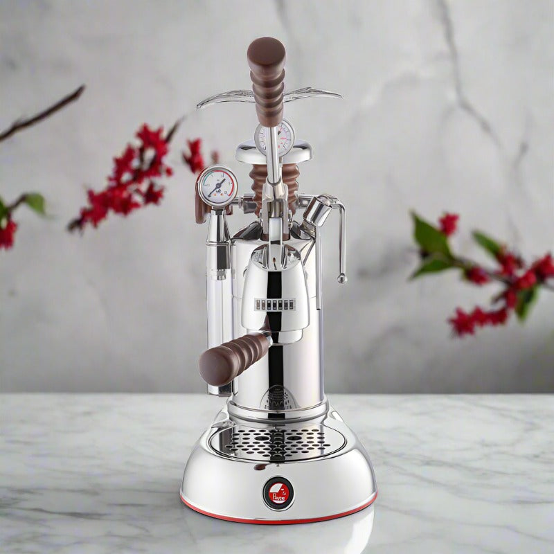 La Pavoni Esperto Abile Coffee Machine in polished chrome with wooden accents, featuring dual pressure gauges, lever-operated brewing system, and drip tray, displayed on marble surface with red floral decorations in background
