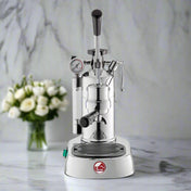 La Pavoni Professional Lusso coffee machine in chrome finish with manual lever, pressure gauge, and steam wand, displayed on marble countertop. Elegant design features polished metal construction and classic Italian espresso maker styling.