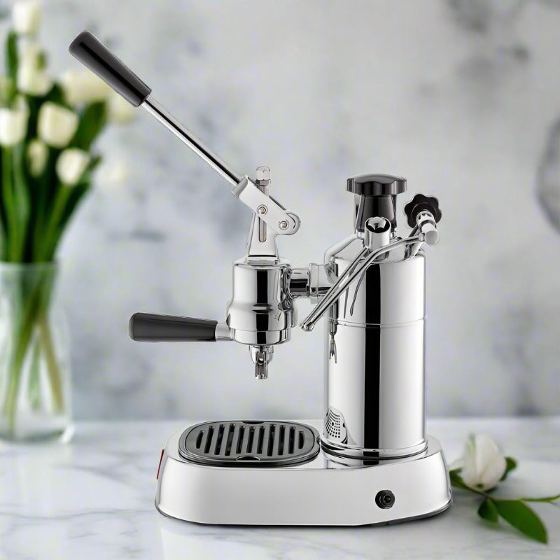 La Pavoni Professional Lusso coffee machine in polished chrome finish with manual lever, steam wand, and drip tray, displayed on marble countertop with white tulips in background, showcasing its classic Italian design and professional-grade espresso-making capabilities.