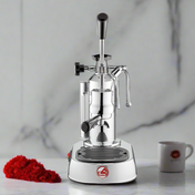La Pavoni Europiccola Lusso manual lever espresso machine in polished chrome finish with white base, displayed alongside a red table decoration and coffee cup against marble background