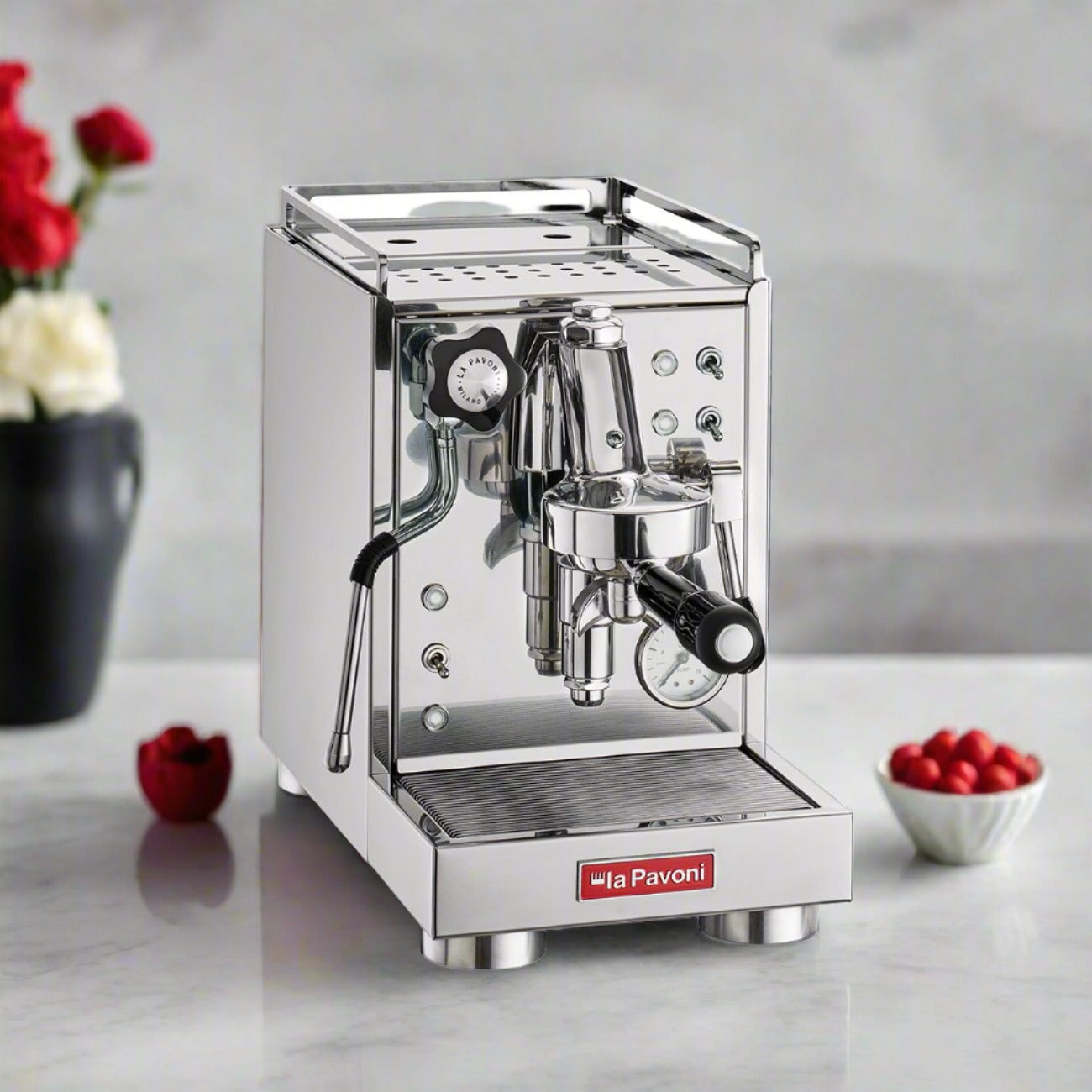 La Pavoni Cellini Mini Coffee Machine in polished stainless steel, featuring professional E61 group head, steam wand, and pressure gauge on white countertop with decorative red roses and raspberries in background